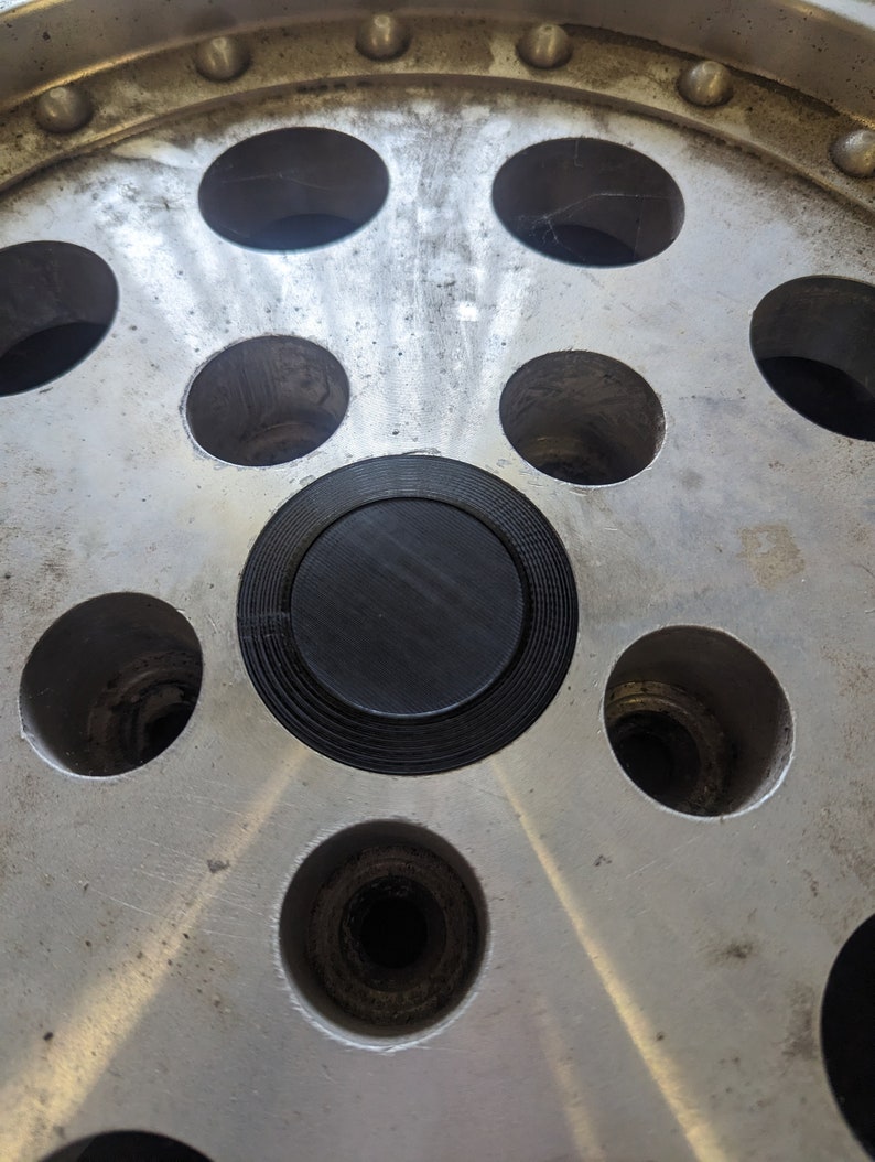 May include: A close-up of a silver car wheel with a black center cap. The wheel has a series of evenly spaced holes around the rim.