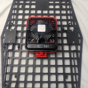 May include: Black, grid-patterned mounting panel with a red and black portable fan attached. The fan has a black blade guard and a red frame. The panel has several mounting brackets and hooks.
