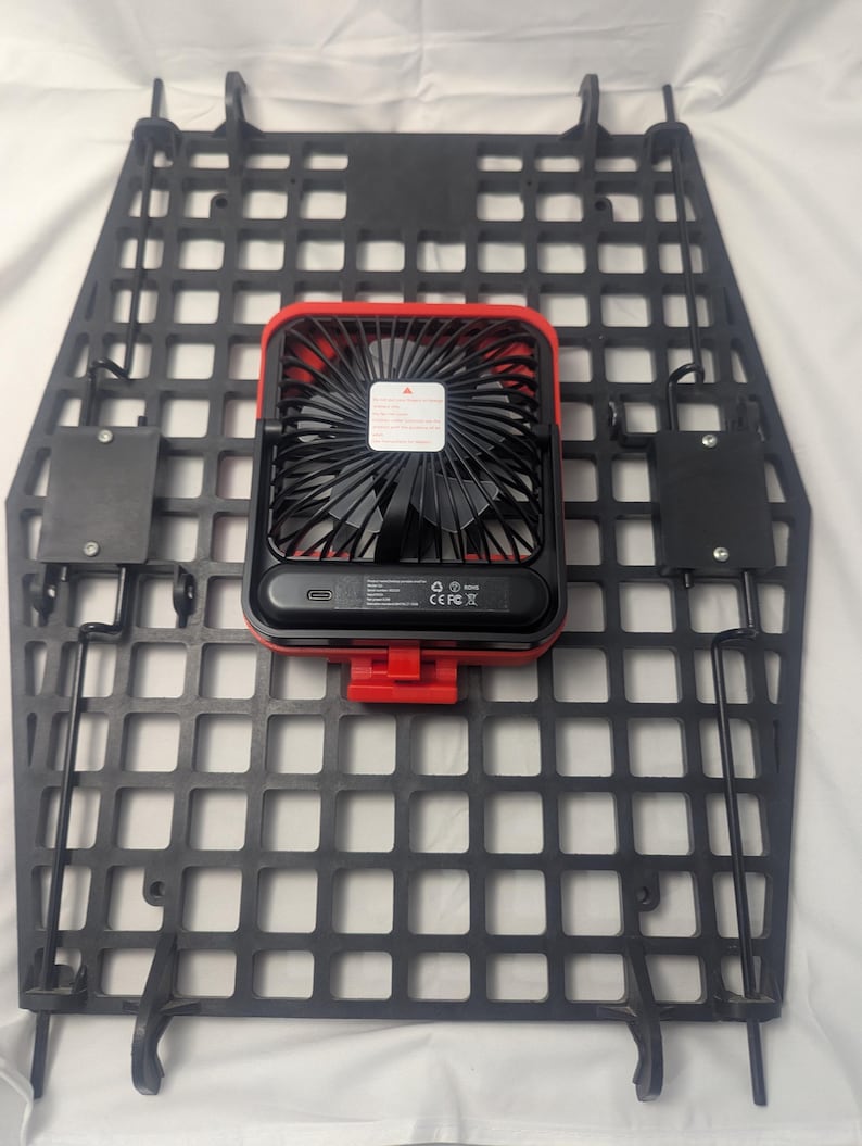May include: Black, grid-patterned mounting panel with a red and black portable fan attached. The fan has a black blade guard and a red frame. The panel has several mounting brackets and hooks.
