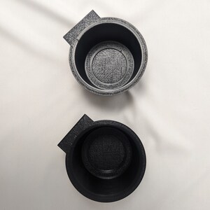 May include: Two black plastic cup holders with a textured surface. The cup holders have a small lip on one side.