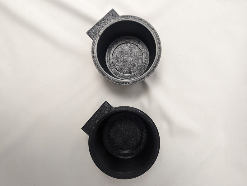May include: Two black plastic cup holders with a textured surface. The cup holders have a small lip on one side.