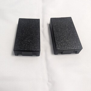 May include: Two black rectangular blocks with a textured surface are displayed against a white background. The blocks are slightly angled, with a small notch at the bottom. The blocks appear to be made of a durable material.