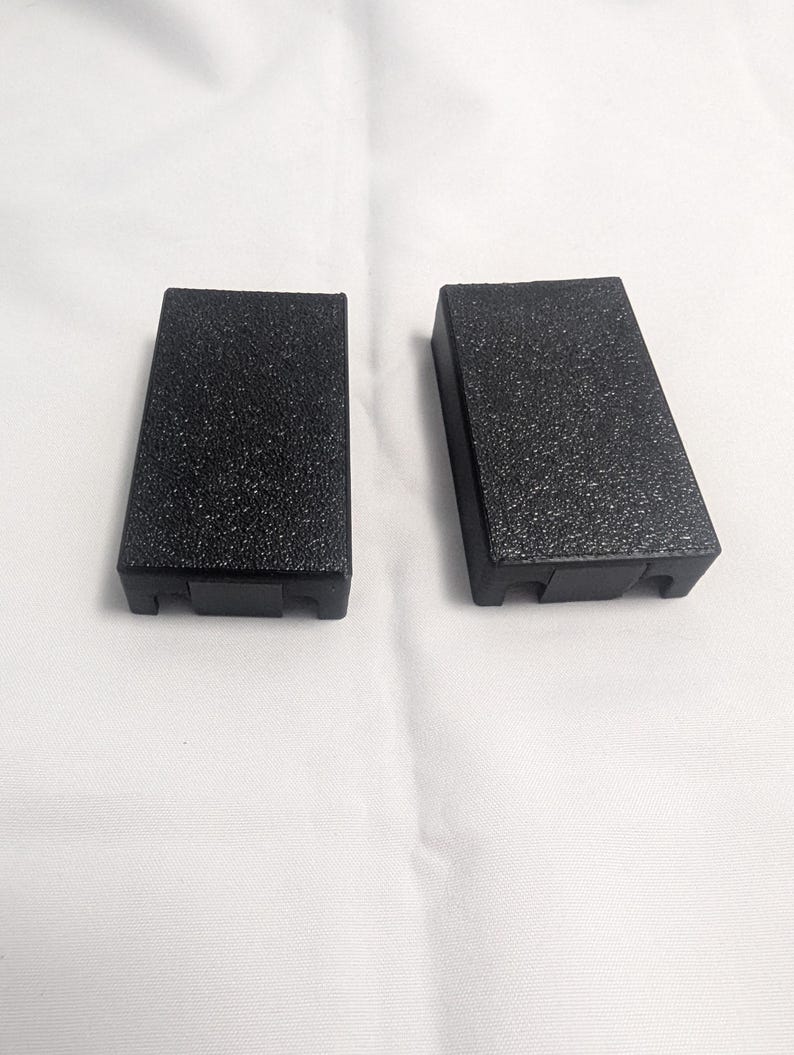 May include: Two black rectangular blocks with a textured surface are displayed against a white background. The blocks are slightly angled, with a small notch at the bottom. The blocks appear to be made of a durable material.