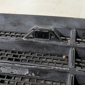 May include: Close-up of a black plastic grille with a small black sensor in the centre. The sensor is rectangular and has a small opening in the centre.