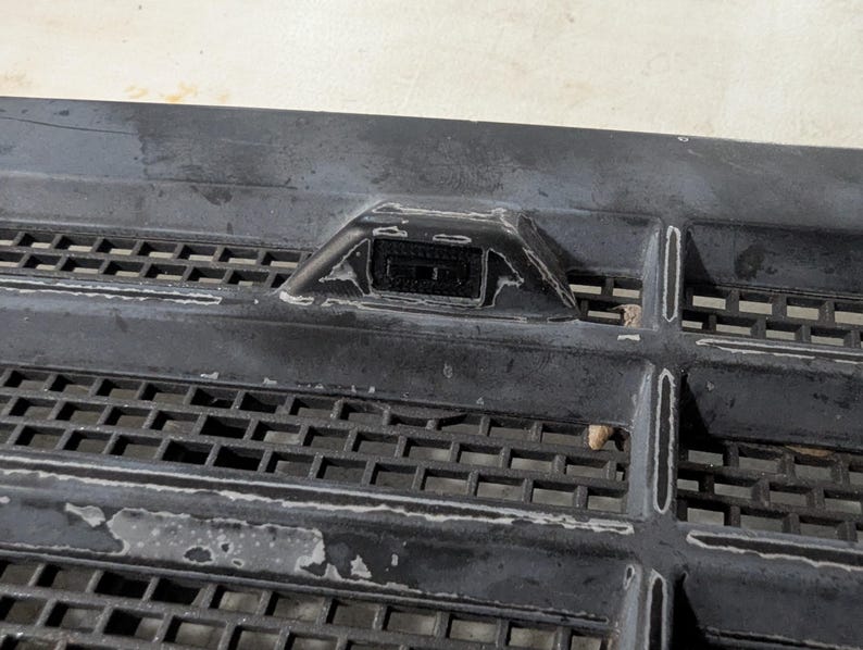 May include: Close-up of a black plastic grille with a small black sensor in the center. The sensor is rectangular and has a small opening in the center.