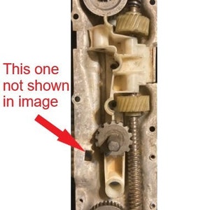 May include: Close-up of a metal gear mechanism with multiple gears and a spring. The mechanism is mounted on a metal plate with several holes. There are three red arrows pointing to different parts of the mechanism.