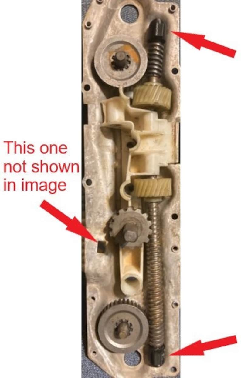 May include: Close-up of a metal gear mechanism with multiple gears and a spring. The mechanism is mounted on a metal plate with several holes. There are three red arrows pointing to different parts of the mechanism.
