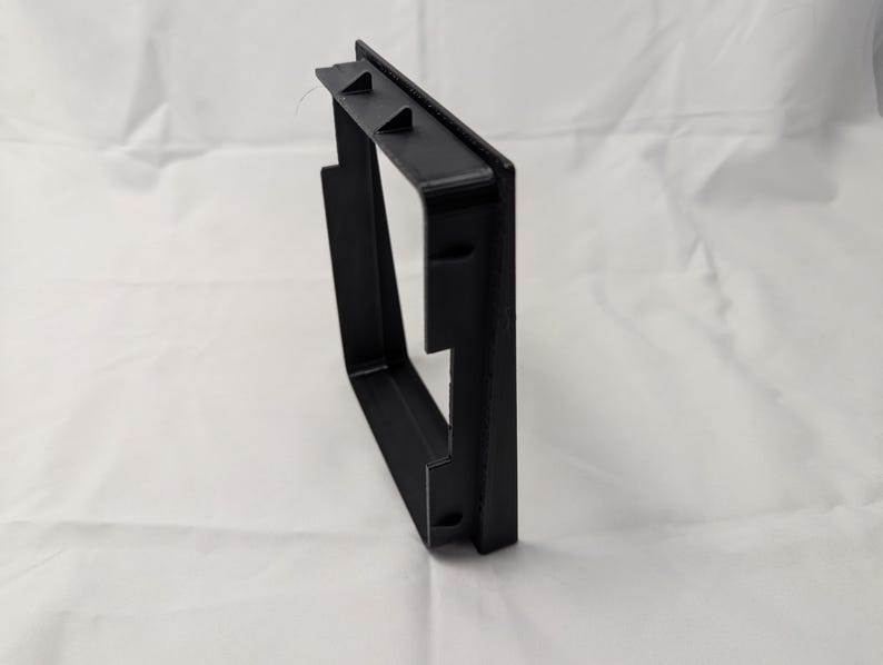 May include: Black plastic frame with a square opening. The frame is designed to be mounted on a wall.
