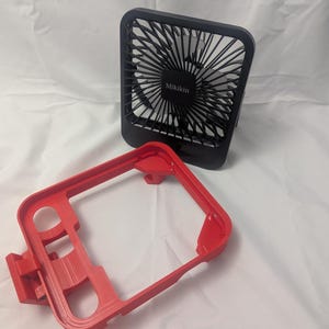 May include: A black Mikikin brand portable fan with a red frame. The fan has a square shape with a protective grill and a series of blades. The red frame has a rectangular shape with several openings.