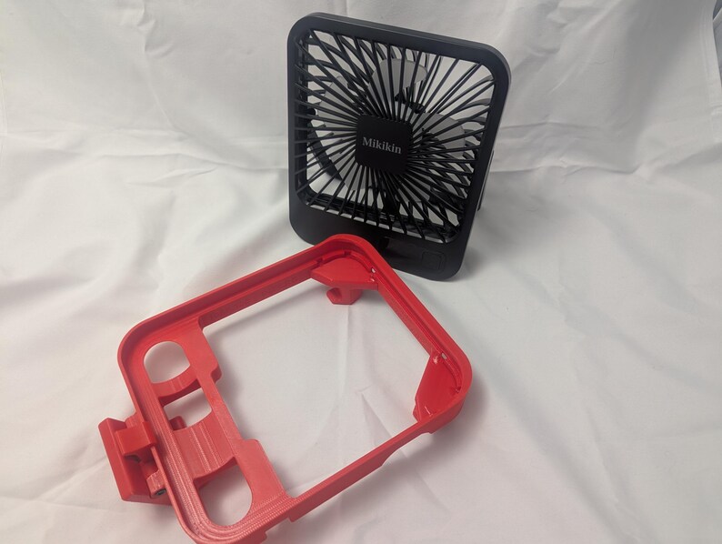 May include: A black Mikikin brand portable fan with a red frame. The fan has a square shape with a protective grill and a series of blades. The red frame has a rectangular shape with several openings.
