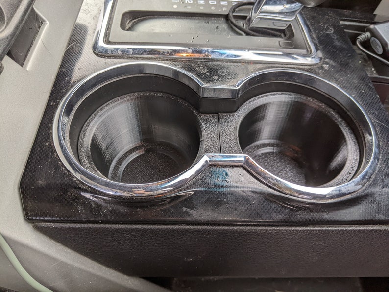 May include: Close-up of a black car interior with two chrome-rimmed cup holders. The cup holders are empty.