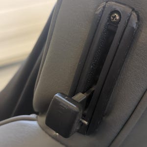 May include: A black plastic lever with a gray fabric background. The lever is attached to a gray fabric seat.