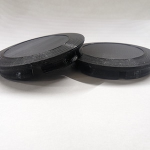 May include: Two black plastic round covers with a textured surface and a raised lip around the edge.