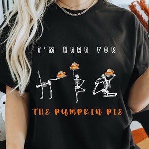 Könnte beinhalten: Schwarzes T-Shirt mit einem Motiv von Skeletten, die Kürbiskuchen halten, und dem Text "Ich bin hier für den Kürbiskuchen".