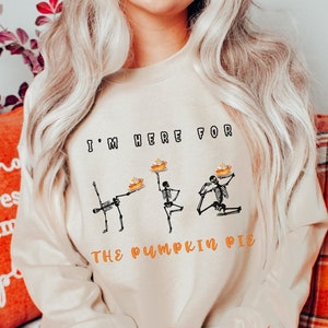 Op de afbeelding: Een beige sweatshirt met de tekst "I'm here for the pumpkin pie" en een afbeelding van drie skeletten die pompoentaarten vasthouden.