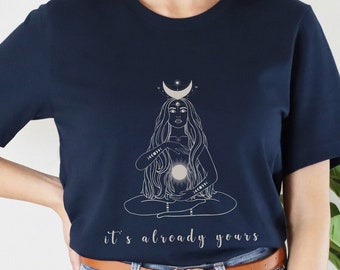 May include: A navy blue t-shirt with a white graphic of a woman with long hair holding a glowing orb. The graphic is surrounded by stars and a crescent moon. The text "it's already yours" is printed below the graphic.