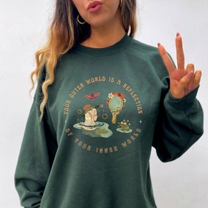 May include: A dark green sweatshirt with a graphic print of a woman with a butterfly and a mirror. The text on the sweatshirt reads "Your outer world is a reflection of your inner world."