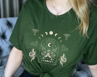 Può includere: Una maglietta verde con una stampa grafica di una falena, serpenti, rane e lune. La grafica è di colore beige chiaro.