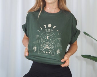 Può includere: Una maglietta verde scuro con una stampa grafica di una falena, fasi lunari e il numero 222. La grafica è di colore crema.