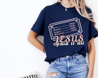 Könnte beinhalten: Marineblaues T-Shirt mit dem Aufdruck "JESUS paid it all". Das Design zeigt ein Ticket mit den Wörtern "Sins, Shame, Anger, Mistakes" und "PAID". Das Shirt wird mit blauen Jeans kombiniert.