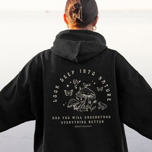 Puede incluir: Sudadera con capucha negra con un estampado gráfico blanco de setas y mariposas. El texto "Mira profundamente en la naturaleza y entenderás todo mejor" está impreso debajo del gráfico.