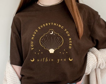 May include: A brown t-shirt with a gold graphic that says "You have everything you need within you". The graphic features a crescent moon, a sun, and a stylized shell.