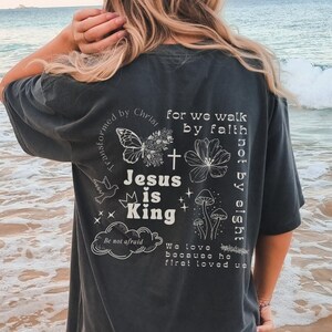 K&ouml;nnte beinhalten: Ein dunkelgraues T-Shirt mit einem wei&szlig;en Grafikdruck, der einen Schmetterling, Blumen, Pilze und den Text "Transformed by Christ", "For we walk by faith, not by sight", "Jesus is King", "Be not afraid", "We love because he first loved us" zeigt.