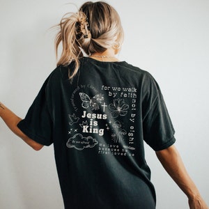 K&ouml;nnte beinhalten: Schwarzes T-Shirt mit einem wei&szlig;en Grafikdesign mit einem Schmetterling, einem Kreuz, einer Krone und dem Text "Jesus is King". Der Text "for we walk by faith not by sight" ist ebenfalls auf dem Shirt enthalten.