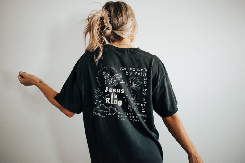 K&ouml;nnte beinhalten: Schwarzes T-Shirt mit einem wei&szlig;en Grafikdesign mit einem Schmetterling, einem Kreuz, einer Krone und dem Text "Jesus is King". Der Text "for we walk by faith not by sight" ist ebenfalls auf dem Shirt enthalten.
