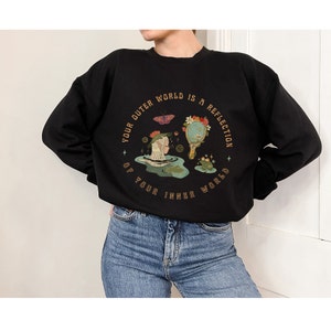 May include: Black sweatshirt with a graphic print of a woman and a frog in a pond. The text around the graphic reads "Your outer world is a reflection of your inner world."