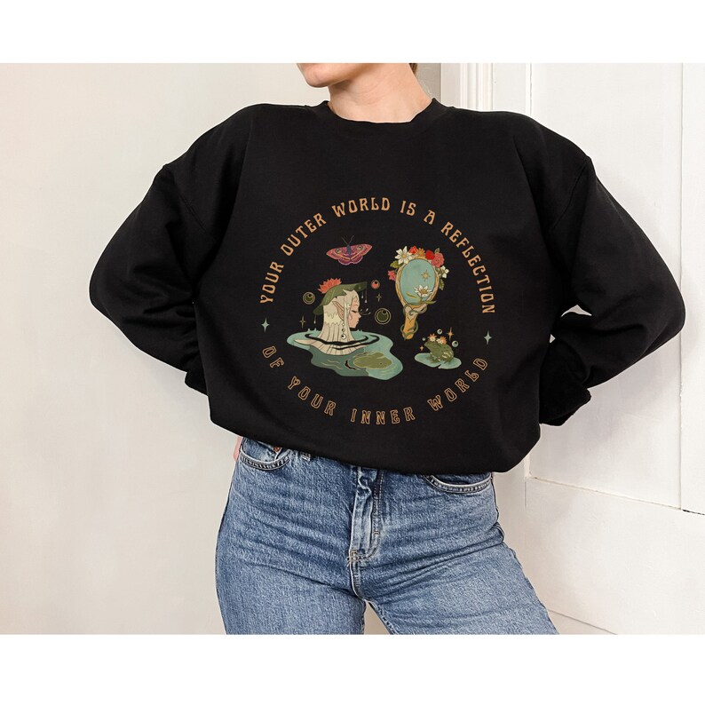 May include: Black sweatshirt with a graphic print of a woman and a frog in a pond. The text around the graphic reads "Your outer world is a reflection of your inner world."