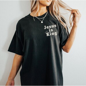 K&ouml;nnte beinhalten: Ein schwarzes T-Shirt mit dem Aufdruck "Jesus is King" auf der Vorderseite. Der Text ist in einer wei&szlig;en, Retro-Schriftart mit einer kleinen Krone &uuml;ber dem "i".