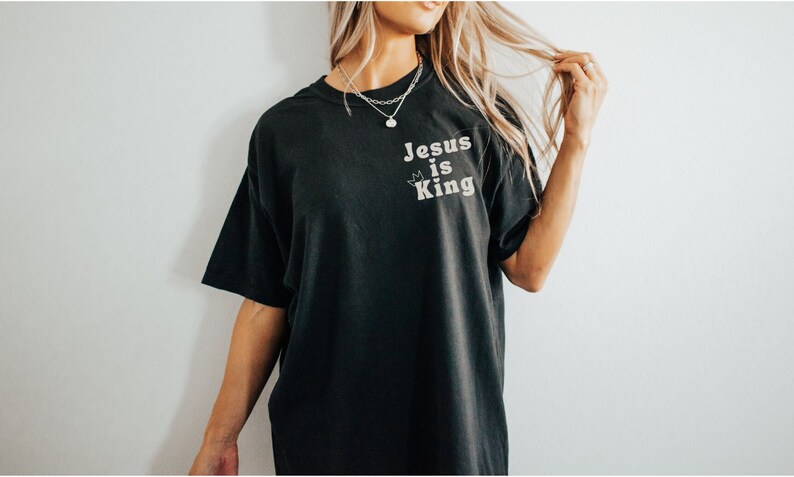 K&ouml;nnte beinhalten: Ein schwarzes T-Shirt mit dem Aufdruck "Jesus is King" auf der Vorderseite. Der Text ist in einer wei&szlig;en, Retro-Schriftart mit einer kleinen Krone &uuml;ber dem "i".