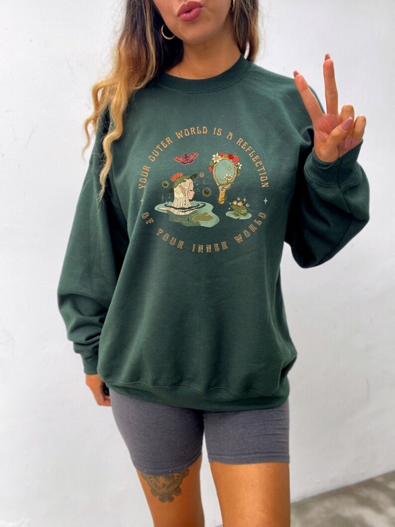 May include: A dark green sweatshirt with a graphic print of a woman with a butterfly and a mirror. The text on the sweatshirt reads "Your outer world is a reflection of your inner world."