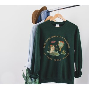 May include: A dark green sweatshirt with a graphic print of a woman in a pond with a frog and a butterfly. The text on the sweatshirt reads "Your outer world is a reflection of your inner world."