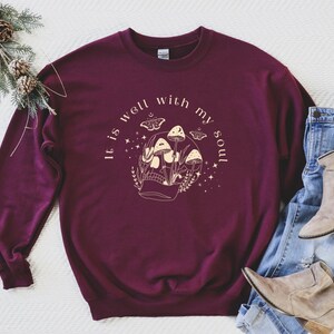 May include: Burgundy sweatshirt with a graphic of a skull with mushrooms growing out of it. The text "It is well with my soul" is printed around the graphic.