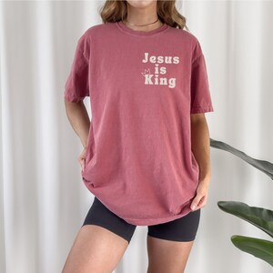 K&ouml;nnte beinhalten: Ein &uuml;berdimensioniertes T-Shirt in Marone mit dem Text "Jesus is King" in Wei&szlig; mit einer Krone &uuml;ber dem Wort "is".