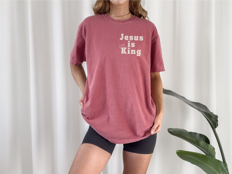 K&ouml;nnte beinhalten: Ein &uuml;berdimensioniertes T-Shirt in Marone mit dem Text "Jesus is King" in Wei&szlig; mit einer Krone &uuml;ber dem Wort "is".