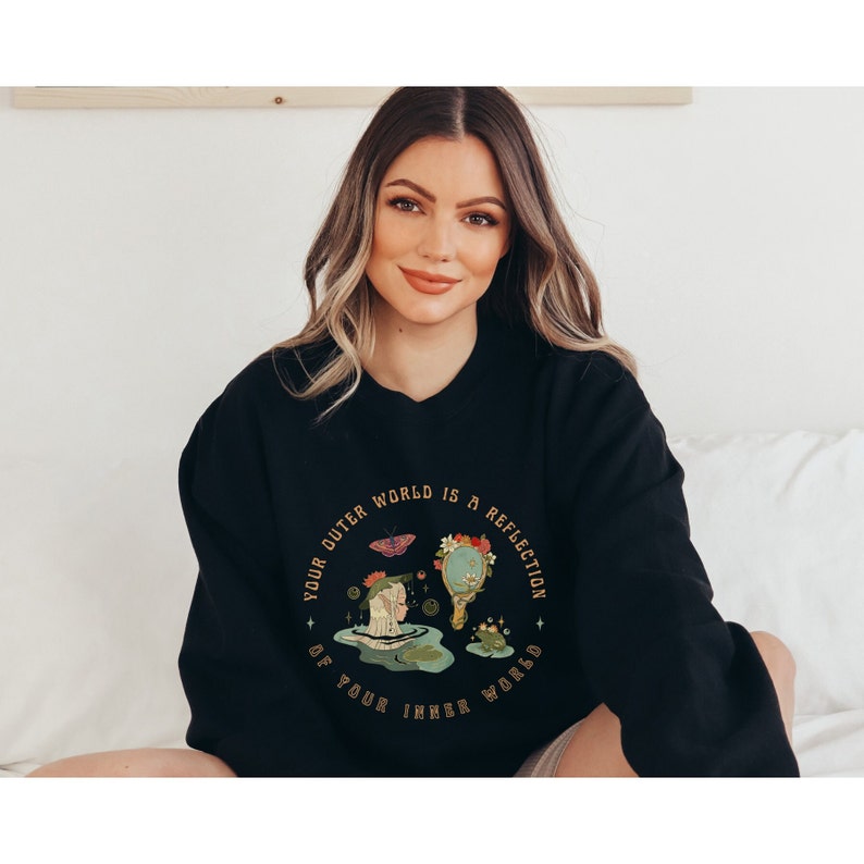 May include: Black sweatshirt with a graphic print of a woman in a pond with a butterfly, a mirror, and a frog. The text on the graphic reads "Your outer world is a reflection of your inner world."