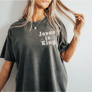 K&ouml;nnte beinhalten: Ein dunkelgraues T-Shirt mit dem Text "Jesus is King" in wei&szlig;en Buchstaben. Der Text ist mittig auf dem Shirt platziert und von zwei kleinen Kronen umgeben.