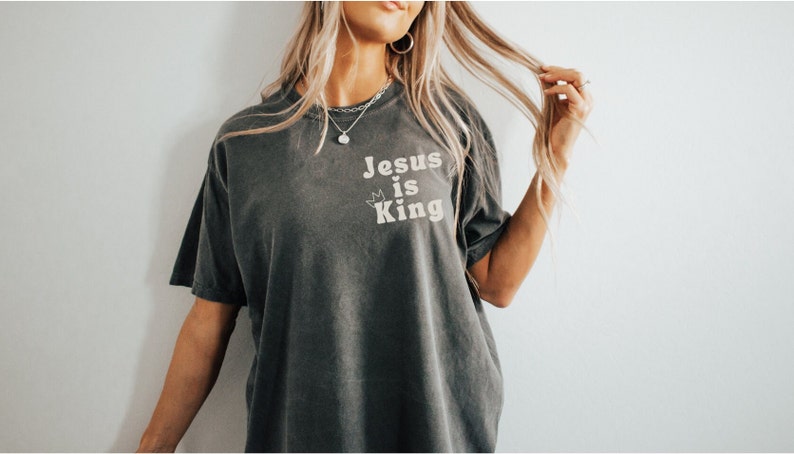 K&ouml;nnte beinhalten: Ein dunkelgraues T-Shirt mit dem Text "Jesus is King" in wei&szlig;en Buchstaben. Der Text ist mittig auf dem Shirt platziert und von zwei kleinen Kronen umgeben.