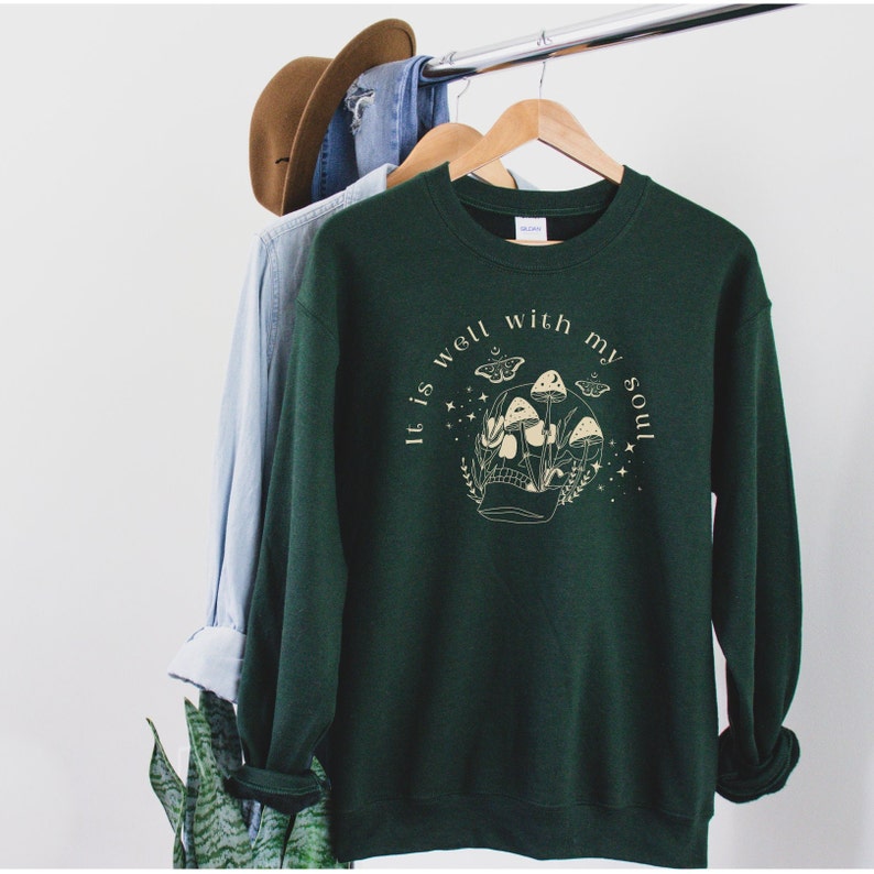 May include: A dark green sweatshirt with a white graphic print of a skull with mushrooms growing out of it. The text "It is well with my soul" is printed around the skull.