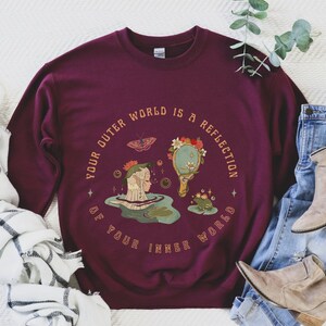 May include: Burgundy sweatshirt with a graphic design of a woman looking into a mirror reflecting a forest scene. The text reads "Your outer world is a reflection of your inner world."