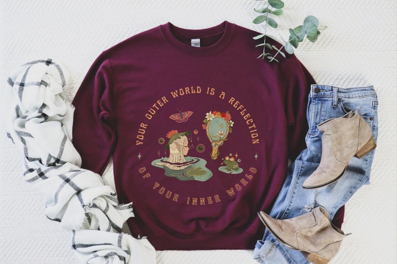 May include: Burgundy sweatshirt with a graphic design of a woman looking into a mirror reflecting a forest scene. The text reads "Your outer world is a reflection of your inner world."