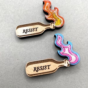Puede incluir: Dos pines de madera con forma de botella con la palabra "RESIST" en letras negras. Un pin se combina con un pin en forma de llama en naranja, amarillo y blanco. El otro pin de botella se combina con un pin en forma de llama en rosa, azul y blanco.