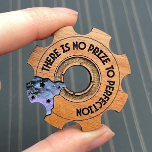 May include: A gear-shaped wooden token featuring the engraved text THERE IS NO PRIZE TO PERFECTION. A section of the gear is replaced by a purple and blue marbled resin inlay decorated with small black star shapes and a central circular cutout.