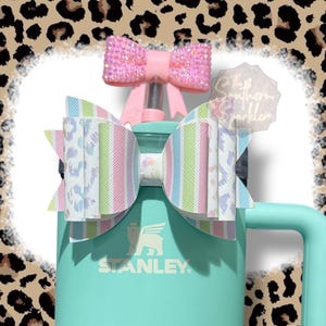 May include: A teal Stanley tumbler with a large pastel striped bow and a smaller pink bow. The bows are attached to the lid. The text "The Southern Sparkler" is visible on a clear tag.