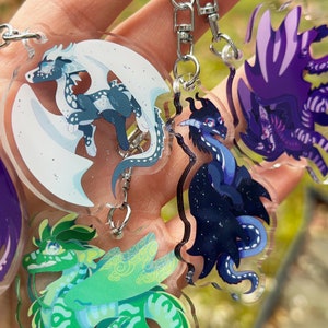 May include: Five colourful dragon keychains with different designs. The dragons are made of clear acrylic with printed details. The keychains are attached to silver chains.