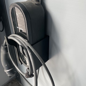 Könnte beinhalten: Eine schwarze Ladestation für Elektrofahrzeuge mit einem aufgerollten Kabel. Die Station hat ein digitales Display mit dem Text "e-tron" darauf.