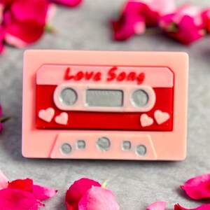 May include: A pink cassette tape with a red center and the words "Love Song" in white. The cassette has two hearts on the bottom right and left corners. The background is a blurred gray surface with pink flower petals.